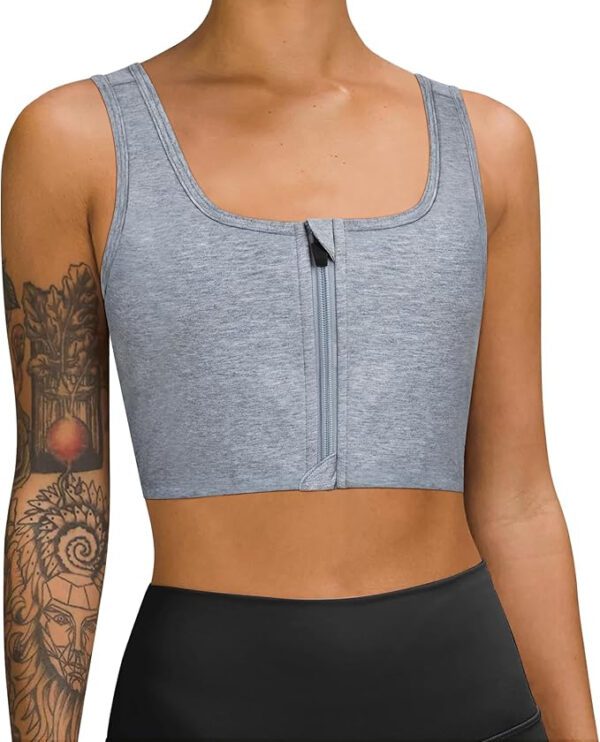 A person wearing a light gray, ftm chest binder with a front zipper and high-waisted leggings. The person's right arm has intricate tattoos, including designs of faces and geometric patterns. The background is white.