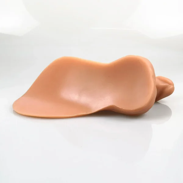 A smooth, flesh-toned silicone p*nis prosthetic lies on a white surface, showcasing intricate detailing and curved edges in subtle beige hues.