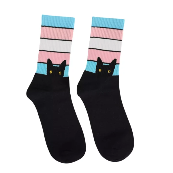 A pair of trans flag socks with colorful stripes at the top in blue, pink, and white, representative of the trans pride flag. The bottom part of the socks is black, featuring a design of a black cat's head with yellow eyes peeking up from the ankle area.