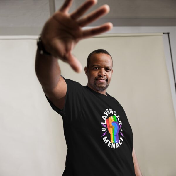 A person stands in a room wearing an Unisex Classic Tee featuring a vibrant fist graphic and the words "Lavender Menace 20XX." They extend their arm towards the camera, partially obscuring their face with their hand.