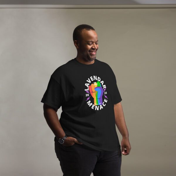 A smiling man dons a black Unisex Classic Tee with bold, rainbow-colored "Lavender Menace" graphics and "2024" emblazoned on each side, all set against a simple backdrop.